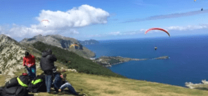 Los parapentistas sobrevuelan el paisaje costero de la Costa Cántabra, con colinas y mar bajo un cielo parcialmente nublado. Una pareja planea junta usando "Vuela en Pareja sobre la Costa Cántabra", armonizando con el viento. - ForTwo - Planes para parejas