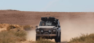 Un vehículo de Aventura en 4x4 por Girona se embarca en un viaje, navegando por un camino de tierra polvoriento en el vasto paisaje desértico bajo un cielo azul claro. - ForTwo - Planes para parejas