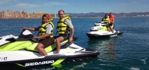 Dos parejas disfrutan de una Aventura en Moto de Agua en Valencia, surcando el agua a toda velocidad bajo el sol. Con chalecos salvavidas puestos, se encuentran rodeados por el hermoso paisaje de los edificios de Valencia y las montañas lejanas. - ForTwo - Planes para parejas