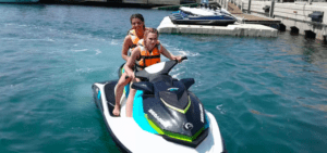 Dos personas con chalecos salvavidas disfrutan de una Aventura en Moto de Agua en Valencia, navegando en una moto acuática por aguas cristalinas cerca de un muelle. - ForTwo - Planes para parejas