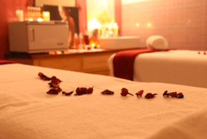 Una sala de spa poco iluminada que recuerda el ambiente tranquilo de los Baños Árabes de Menorca, adornada con pétalos de rosa en una mesa de masajes y velas suavemente encendidas en el fondo. - ForTwo - Planes para parejas