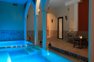 Una piscina cubierta con iluminación azul, columnas arqueadas que recuerdan a los Baños Árabes de Menorca, paredes de azulejos, faroles decorativos y una pequeña chimenea crean un ambiente sereno. - ForTwo - Planes para parejas