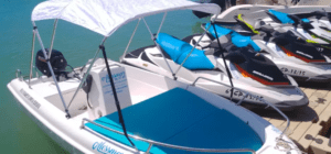 Un barco sin licencia en Alcossebre, con casco blanco, toldo y asientos azules, está atracado junto a motos de agua en las aguas turquesas y disponible para alquilar. - ForTwo - Planes para parejas