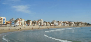 La playa de arena de Alcossebre cuenta con olas suaves y edificios vibrantes bajo un cielo azul claro, mientras que un "Barco sin licencia en Alcossebre" se mece suavemente en el agua brillante cercana. - ForTwo - Planes para parejas