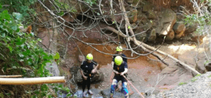 Tres individuos con cascos y equipo de escalada exploran una zona boscosa rocosa con un arroyo, similar a la aventura del "Barranquismo en la Sierra de Cardeña y Montoro". - ForTwo - Planes para parejas