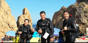 Tres buceadores, emocionados por su Bautismo de Buceo en Tossa de Mar, posan en una playa rocosa con su equipo, sonriendo contra el telón de fondo del cielo azul de la ciudad y los majestuosos acantilados. - ForTwo - Planes para parejas
