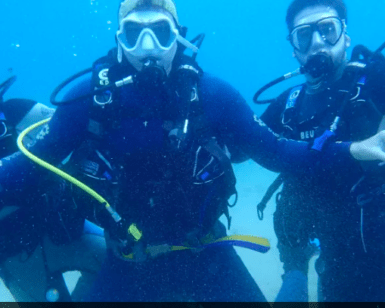 Tres buceadores bajo el agua en Tossa de Mar, con trajes de neopreno y máscaras negras, hacen un gesto a la cámara durante su Bautismo de Buceo en Tossa de Mar. - ForTwo - Planes para parejas