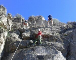 Bajo un cielo azul claro, tres escaladores escalan un acantilado rocoso, asegurados con cuerdas y equipo, abrazando plenamente "Bautismo de escalada en Valencia: ¡una experiencia llena de desafío y conexión!. - ForTwo - Planes para parejas