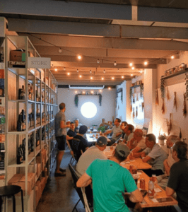En una acogedora cafetería de Valencia, los clientes conversan enfrascados en una mesa rodeada de estanterías llenas de botellas. Disfrutan de una Cata de Cervezas Artesanas de Valencia, saboreando los sabores únicos de diferentes cervezas artesanales. - ForTwo - Planes para parejas