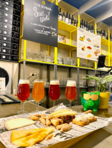 En el mostrador, junto a una rana verde, se encuentran una selección de cervezas ácidas y una tabla de quesos. Detrás, un menú presenta la experiencia "Cata de Cervezas Artesanas en Valencia". - ForTwo - Planes para parejas