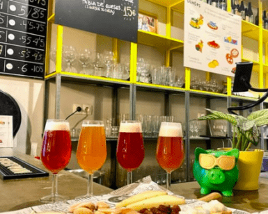En el mostrador, junto a una rana verde, se encuentran una selección de cervezas ácidas y una tabla de quesos. Detrás, un menú presenta la experiencia "Cata de Cervezas Artesanas en Valencia". - ForTwo - Planes para parejas