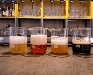 Cuatro vasos de Cervezas Artesanas en tonos vibrantes se encuentran en una barra de bar, con estantes y cristalería brillante detrás: una escena acogedora para una experiencia de Cata de Cervezas Artesanas en Valencia. - ForTwo - Planes para parejas