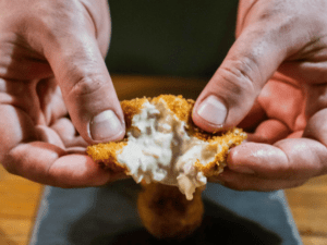 Manos que abren una croqueta crujiente para revelar un relleno cremoso: experimente esta delicia clásica a través de nuestra clase de cocina española en Valencia: tapas y vinos. - ForTwo - Planes para parejas