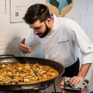 Un chef con bata blanca inspecciona una gran cacerola de paella durante el evento "Clase de cocina española en Valencia: tapas y vinos", que se desarrolla en el interior. - ForTwo - Planes para parejas