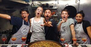 Cinco personas emocionadas con delantales posan en una cocina con Clase de paella tradicional y cena en Valencia y vasos de sangría. - ForTwo - Planes para parejas