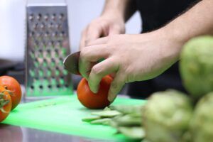 Una persona está cortando un tomate en una tabla de cortar verde cerca de un rallador y alcachofas, preparando ingredientes frescos para una cena auténtica de "Clase de paella tradicional y cena en Valencia". - ForTwo - Planes para parejas