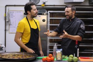 Dos chefs en una cocina valenciana, rodeados de tomates, alcachofas y vino, intercambian consejos para elaborar un plato auténtico durante la Clase de paella tradicional y cena en Valencia. - ForTwo - Planes para parejas