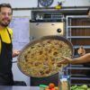 Dos chefs en una cocina sostienen con orgullo una gran sartén de paella vibrante, resaltando los sabores de Valencia, rodeados de verduras frescas y vino, encarnando la esencia de la clase de paella tradicional y cena en Valencia. - ForTwo - Planes para parejas
