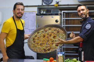 Dos chefs en una cocina sostienen con orgullo una gran sartén de paella vibrante, resaltando los sabores de Valencia, rodeados de verduras frescas y vino, encarnando la esencia de la clase de paella tradicional y cena en Valencia. - ForTwo - Planes para parejas