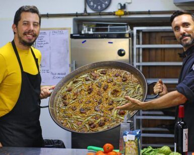 Dos chefs en una cocina sostienen con orgullo una gran sartén de paella vibrante, resaltando los sabores de Valencia, rodeados de verduras frescas y vino, encarnando la esencia de la clase de paella tradicional y cena en Valencia. - ForTwo - Planes para parejas
