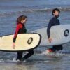 Dos personas en traje de neopreno caminan por el océano, tablas de surf en mano, listas para abrazar las olas después de sus emocionantes Clases de Surf en Valencia. - ForTwo - Planes para parejas