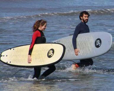 Dos personas en traje de neopreno caminan por el océano, tablas de surf en mano, listas para abrazar las olas después de sus emocionantes Clases de Surf en Valencia. - ForTwo - Planes para parejas