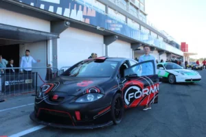 Cerca de los boxes de Valencia, un coche de carreras negro y rojo con el logo de FORM se encuentra, mientras la gente y otros coches se mueven a su alrededor. El ambiente se llena de la emoción del Copilotaje extremo en Valencia. - ForTwo - Planes para parejas