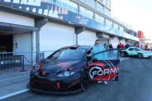Un elegante coche de carreras negro con calcomanías rojas se encuentra frente a la bulliciosa zona de boxes en el circuito de carreras de Valencia, listo para el "Copilotaje extremo en Valencia". - ForTwo - Planes para parejas