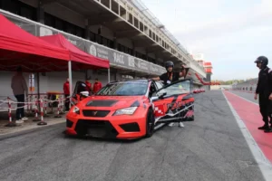 Una persona está parada junto a un auto de carreras rojo en la pista de Valencia, rodeada por un equipo de boxes dedicado, listo para Copilotaje extremo en Valencia. - ForTwo - Planes para parejas