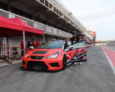 Una persona está parada junto a un auto de carreras rojo en la pista de Valencia, rodeada por un equipo de boxes dedicado, listo para Copilotaje extremo en Valencia. - ForTwo - Planes para parejas