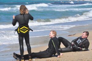 Tres personas en traje de neopreno se relajan y charlan en la playa de arena junto a las olas del mar, tomando un descanso de su Curso de Windsurf en Atalaya Isdabe. - ForTwo - Planes para parejas
