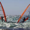 Dos windsurfistas con velas naranjas se enfrentan en el brillante océano cerca de Atalaya Isdabe, perfeccionando sus habilidades bajo un cielo azul claro, mientras participan en el Curso de Windsurf en Atalaya Isdabe. - ForTwo - Planes para parejas
