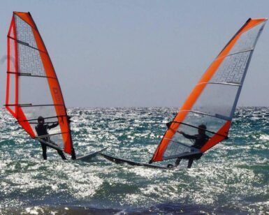 Dos windsurfistas con velas naranjas se enfrentan en el brillante océano cerca de Atalaya Isdabe, perfeccionando sus habilidades bajo un cielo azul claro, mientras participan en el Curso de Windsurf en Atalaya Isdabe. - ForTwo - Planes para parejas