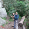 Dos personas sonríen mientras están de pie cerca de una pared de roca, sosteniendo una cuerda de escalada y listas para una emocionante aventura con el "Curso de escalada y rappel en Alange", rodeado de árboles y troncos. - ForTwo - Planes para parejas