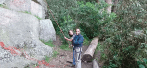 Dos personas sonríen mientras están de pie cerca de una pared de roca, sosteniendo una cuerda de escalada y listas para una emocionante aventura con el "Curso de escalada y rappel en Alange", rodeado de árboles y troncos. - ForTwo - Planes para parejas