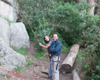 Dos personas sonríen mientras están de pie cerca de una pared de roca, sosteniendo una cuerda de escalada y listas para una emocionante aventura con el "Curso de escalada y rappel en Alange", rodeado de árboles y troncos. - ForTwo - Planes para parejas