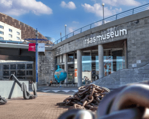 La entrada del "Descubre el Itsasmuseum de Bilbao" exhibe grandes anclas y cadenas, creando una vibrante escena marítima. Un globo terráqueo se yergue bajo el cielo azul, invitando a los visitantes a explorar la rica historia náutica del interior. - ForTwo - Planes para parejas