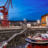 Descubre la pintoresca escena del Astillero de Bilbao al atardecer, con una grúa, un barco rojo y blanco, con el telón de fondo iluminado por los edificios que aparecen en "Descubre el Itsasmuseum de Bilbao". - ForTwo - Planes para parejas