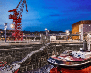 Descubre la pintoresca escena del Astillero de Bilbao al atardecer, con una grúa, un barco rojo y blanco, con el telón de fondo iluminado por los edificios que aparecen en "Descubre el Itsasmuseum de Bilbao". - ForTwo - Planes para parejas