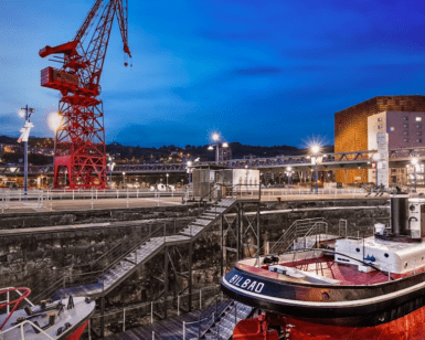 Descubre la pintoresca escena del Astillero de Bilbao al atardecer, con una grúa, un barco rojo y blanco, con el telón de fondo iluminado por los edificios que aparecen en "Descubre el Itsasmuseum de Bilbao". - ForTwo - Planes para parejas