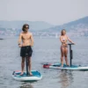 Dos personas sobre tablas de E-Paddle Surf en Marbella se deslizan por un lago tranquilo, rodeado de colinas lejanas y barcos dispersos, que recuerdan un día sereno. - ForTwo - Planes para parejas