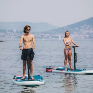 Dos personas sobre tablas de E-Paddle Surf en Marbella se deslizan por un lago tranquilo, rodeado de colinas lejanas y barcos dispersos, que recuerdan un día sereno. - ForTwo - Planes para parejas