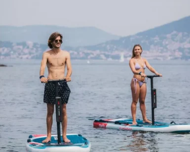 Dos personas sobre tablas de E-Paddle Surf en Marbella se deslizan por un lago tranquilo, rodeado de colinas lejanas y barcos dispersos, que recuerdan un día sereno. - ForTwo - Planes para parejas