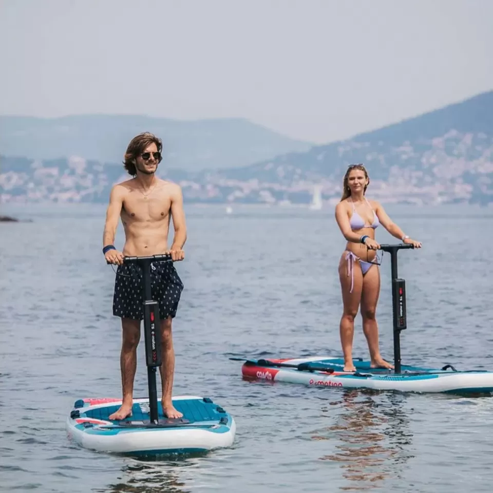 Dos personas sobre tablas de E-Paddle Surf en Marbella se deslizan por un lago tranquilo, rodeado de colinas lejanas y barcos dispersos, que recuerdan un día sereno. - ForTwo - Planes para parejas