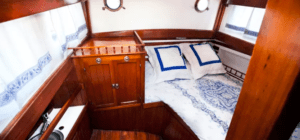 Una acogedora cabaña de madera con una cama cuidadosamente hecha, almohadas y ventanas de ojo de buey refleja el encanto sereno de la Escapada en velero de Ibiza a Formentera. - ForTwo - Planes para parejas