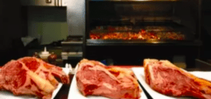 Tres gruesos filetes crudos reposan invitantes en platos ante las brasas de una parrilla, evocando la esencia de una "Escapada gastronómica a Astigarraga" en tu propia cocina. - ForTwo - Planes para parejas