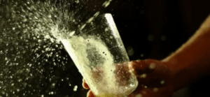 Bajo una iluminación dramática, una mano sostiene un vaso del que salpica un líquido espumoso, evocando la esencia de un brindis con sidra de Escapada gastronómica en Astigarraga. - ForTwo - Planes para parejas