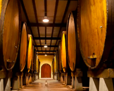 Una bodega poco iluminada con grandes barriles de madera alineados a ambos lados de un estrecho pasillo invita a una escapada gastronómica a Astigarraga. - ForTwo - Planes para parejas