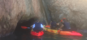 Bajo el radiante sol de Girona, tres aventureros de "Escape Room en Kayak por Girona" reman hacia la entrada de una misteriosa cueva rocosa, ansiosos por afrontar su desafío. - ForTwo - Planes para parejas