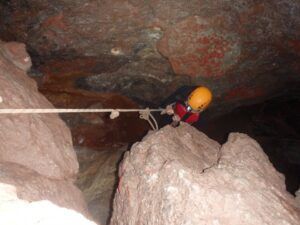 Con un casco, un individuo emprende una aventura extrema escalando paredes rocosas de una cueva usando una cuerda, haciéndose eco del espíritu de "Espeleología acuática en el Túnel del Sumidor: una aventura extrema en pareja". - ForTwo - Planes para parejas